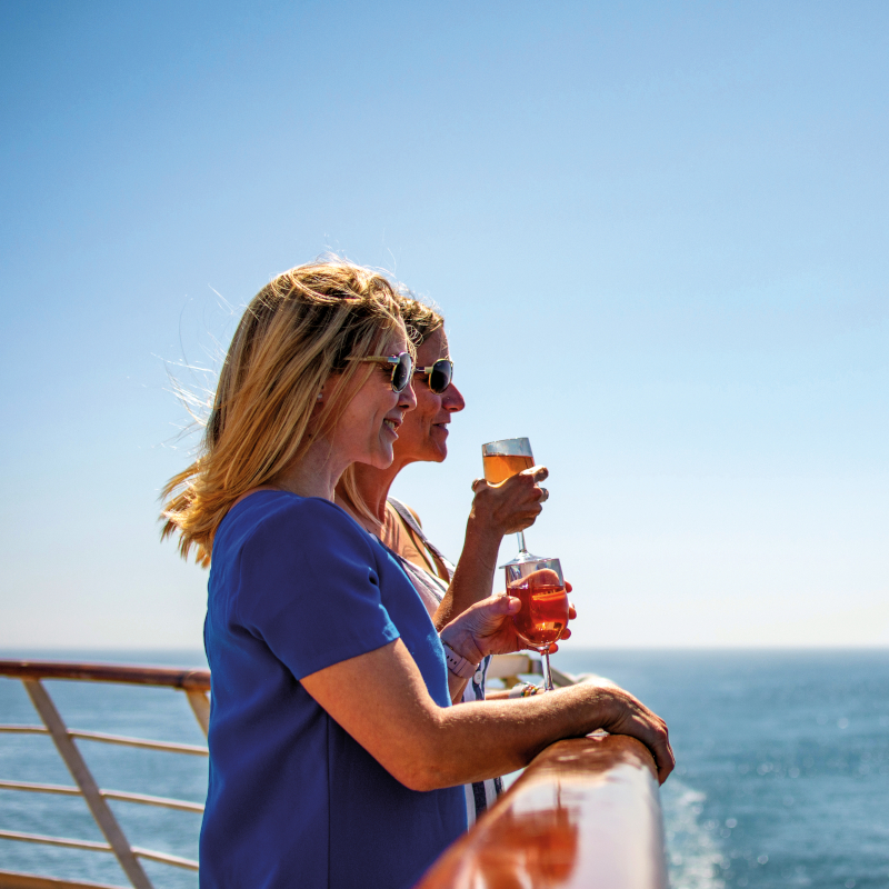 Fred-Olsen-drinks-on-deck Two women standing at the wooden railing of a cruise ship, holding drinks and looking out at a bright blue sea.