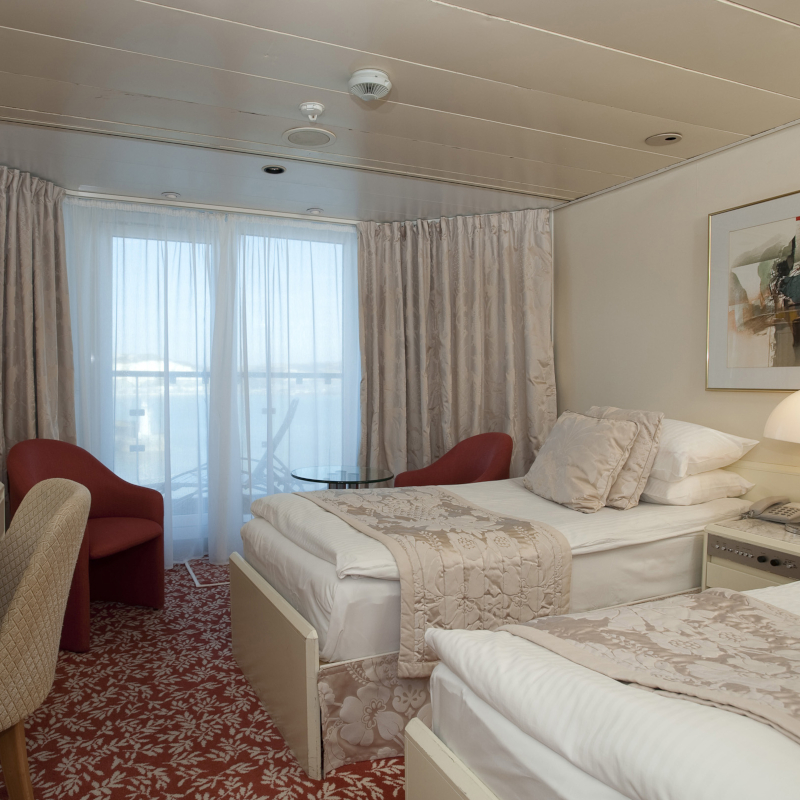 Fred-Olsen-superior-balcony-cabin A cruise ship twin balcony cabin featuring two single beds with beige floral runners, red accent armchairs, and floor-to-ceiling glass doors leading to a balcony.