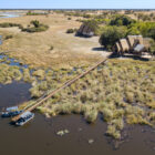 Aerial view of a luxury safari camp with thatched-roof structures connected by a long wooden walkway to a boat dock on a river.