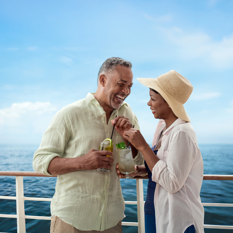Holland-America-couple-on-deck Couple laughing together on a ship deck with tropical cocktails against a sunny blue sky and sea.
