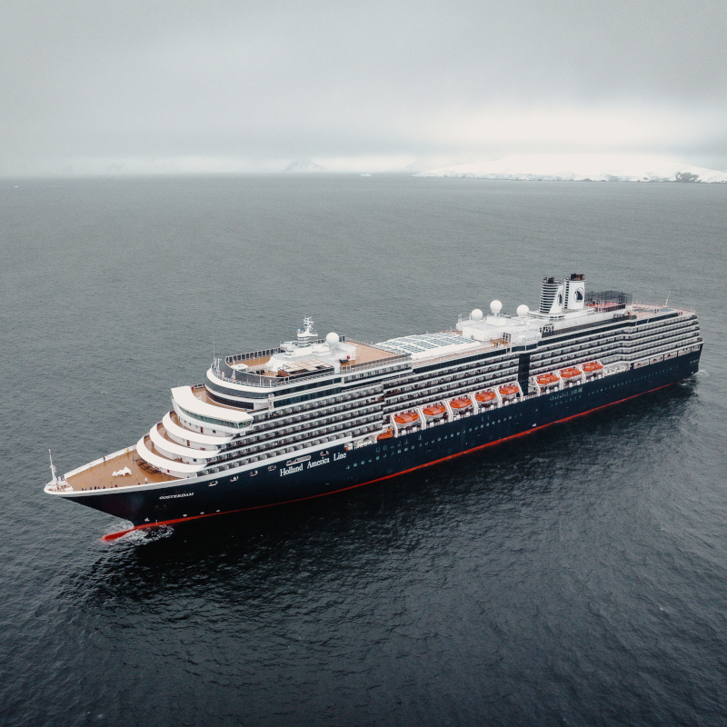 Holland-America-cruise-ship Aerial wide-angle view of a Holland America Line cruise ship navigating calm, deep grey water.