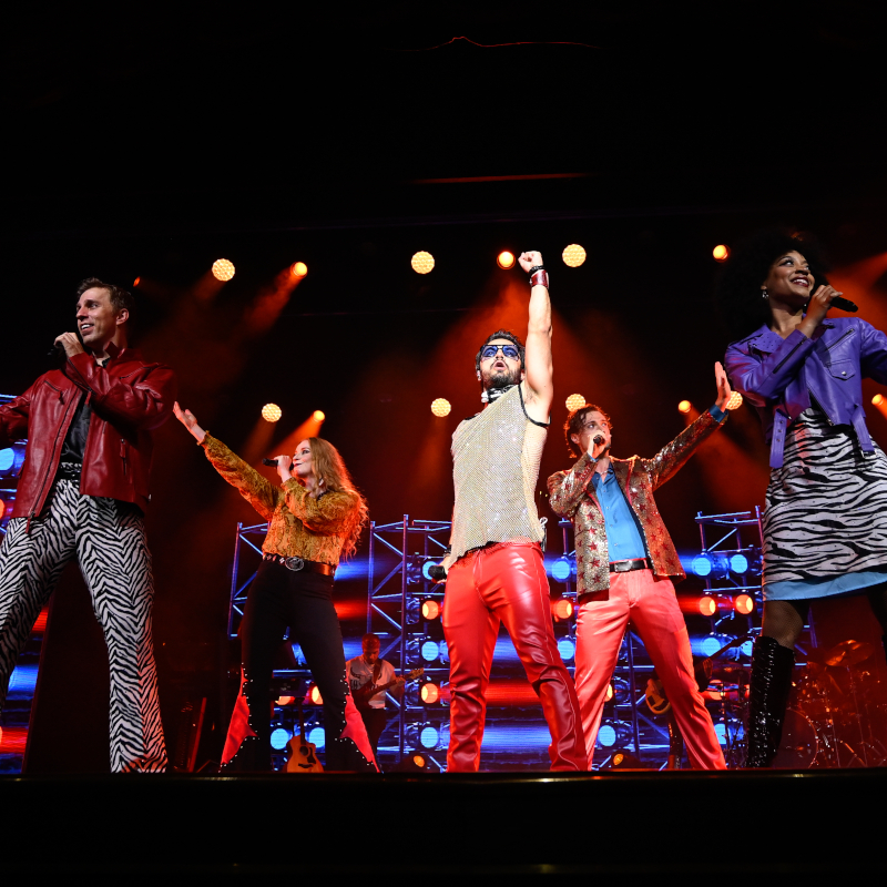 Holland-America-entertainment Five performers in colourful, retro 1970s outfits singing and dancing on a brightly lit stage.