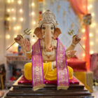 A colourful statue of the Hindu deity Lord Ganesha seated on a platform, adorned with a yellow dhoti, pink and gold sashes, and holding a trident.