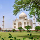 A side view of the white marble Taj Mahal framed by green tree branches in the foreground, with visitors walking along the platform and lawns below.