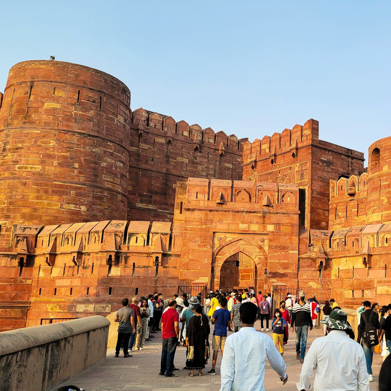 A bustling crowd of tourists walking along a path leading toward the historic, towering red sandstone ramparts and gateways of Agra Fort.