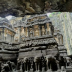 A view from under a rock overhang looking up at the towering, rock-cut Kailasa Temple at Ellora Caves, featuring detailed elephant carvings lining its stone base.