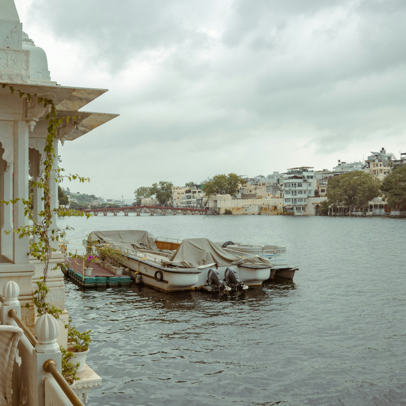 A view of Lake Pichola in Udaipur from a white marble balcony, with two motorboats docked in the water and a distant bridge and city skyline under an overcast sky.