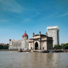 The Gateway of India stone arch standing on the Mumbai waterfront, flanked by the historic Taj Mahal Palace hotel and a modern high-rise.