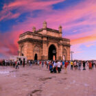 A crowd of people gathered in the stone plaza in front of the Gateway of India arch in Mumbai, under a dramatic pink and purple sunset sky.