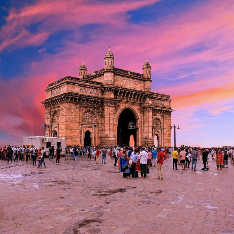 A crowd of people gathered in the stone plaza in front of the Gateway of India arch in Mumbai, under a dramatic pink and purple sunset sky.