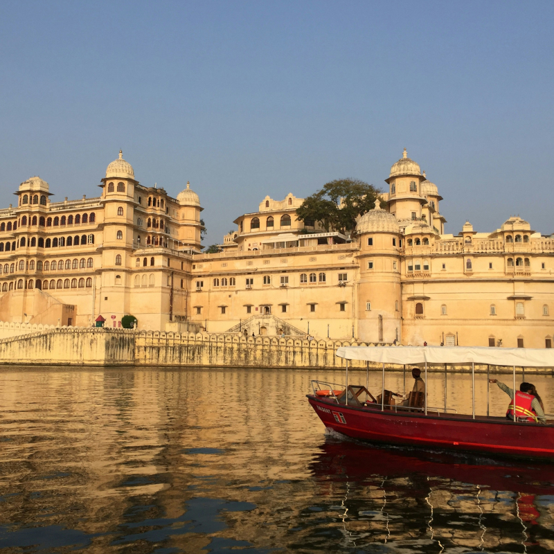 A red and white motorboat carrying passengers on water, with the grand, cream-colored City Palace of Udaipur reflecting in the water in the background.