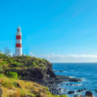 Red and white striped lighthouse on a rocky, grassy cliffside overlooking the ocean under a clear blue sky.