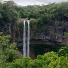 A tall, double-drop waterfall plunging down a steep, dark rocky cliff surrounded by dense tropical green forest.