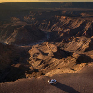 A high-angle, scenic view of a deep, winding rocky canyon landscape with a white off-road vehicle parked on the edge in the foreground.