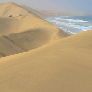 A sweeping view of massive golden sand dunes meeting the ocean waves, with two off-road vehicles driving along a high ridge.