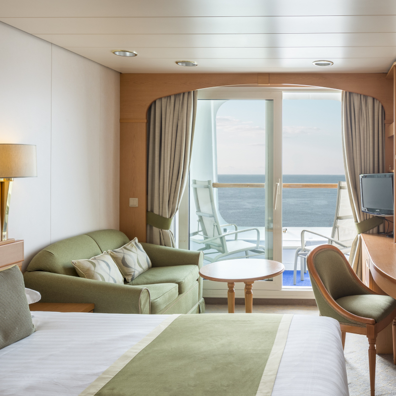 P&O-Cruises-balcony-cabin A modern cruise ship balcony cabin featuring a bed, sofa, and glass sliding door opening to an ocean view.