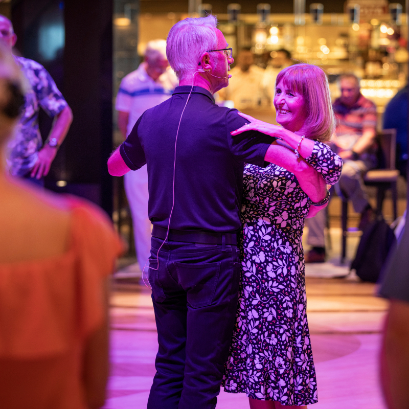 P&O-Cruises-dance-classes A smiling couple enjoying a ballroom dance class under vibrant purple lighting on board.