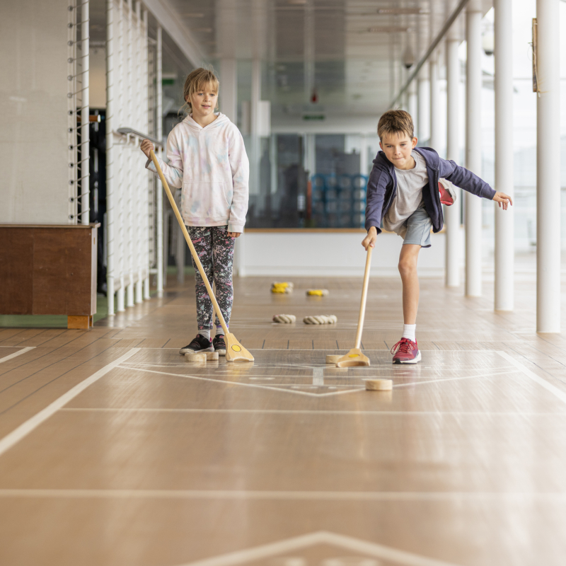 P&O-Cruises-deck-games Two children playing shuffleboard together on the outdoor wooden deck of a cruise ship.