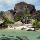 A sandy tropical beach nestled beneath towering, uniquely weathered dark granite boulders and lush palm trees.