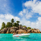 A white speedboat floating in vibrant turquoise water next to a rugged, uniquely shaped rocky island with palm trees.