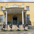 Four bronze historical statues standing on display in front of the white-pillared entrance of a yellow colonial building.