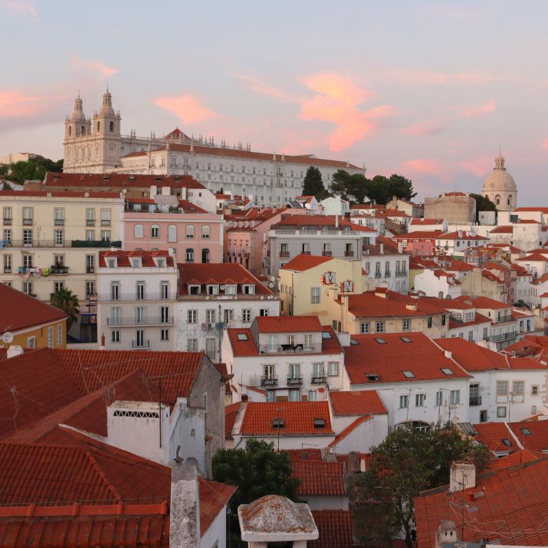 Rooftop view of Lisbon's white buildings with red-tiled roofs under a pink evening sky.