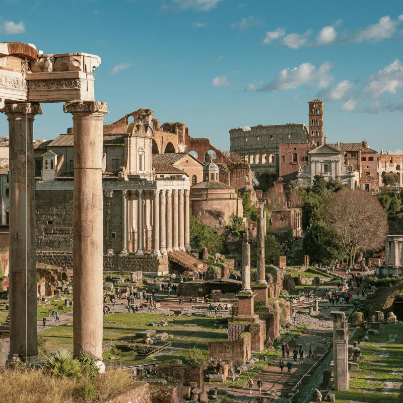 Ruins of the Roman Forum in Rome with columns, ancient stone structures, and the Colosseum in the background.
