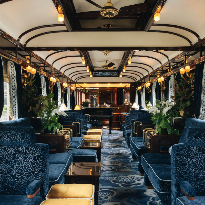 Elegant vintage train bar car with blue plush seating, wooden finishes, and a grand piano at the far end.