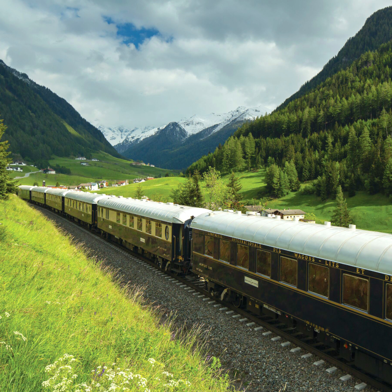 Venice Simplon-Orient-Express train travelling through a green mountain valley with snow-capped peaks.