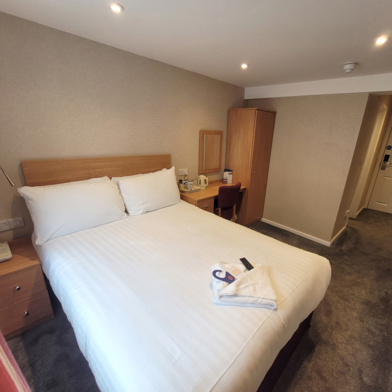 Hotel bedroom featuring a made double bed, wooden furniture, a desk, and grey carpeting.