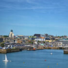 Panoramic harbour view with sailboats on blue water and a coastal city skyline in the background.